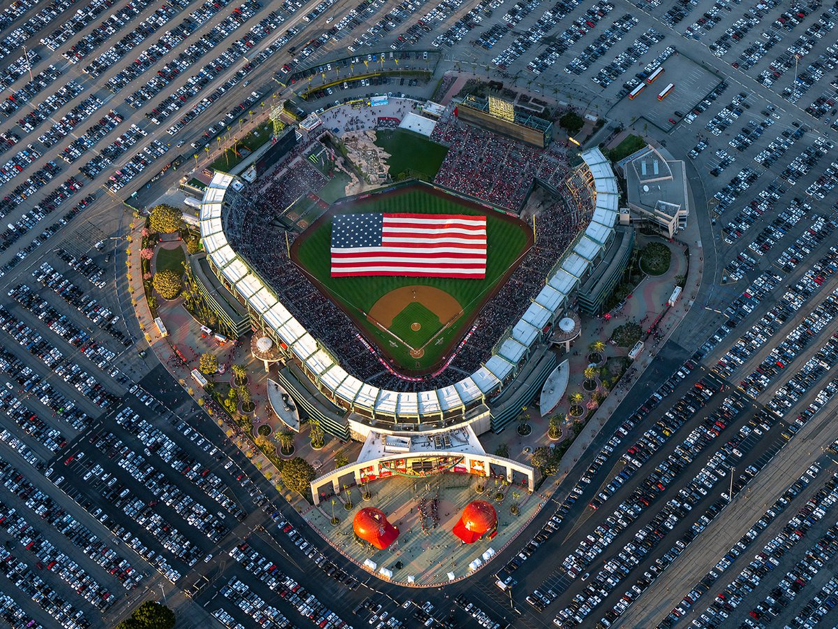 An oblique aerial perspective showcases a grand 300-ft American Flag sprawled across the outfield of Angel Stadium, made possible by 150 active and retired military members, during a rendition of the National Anthem by Jill Marie Burke.