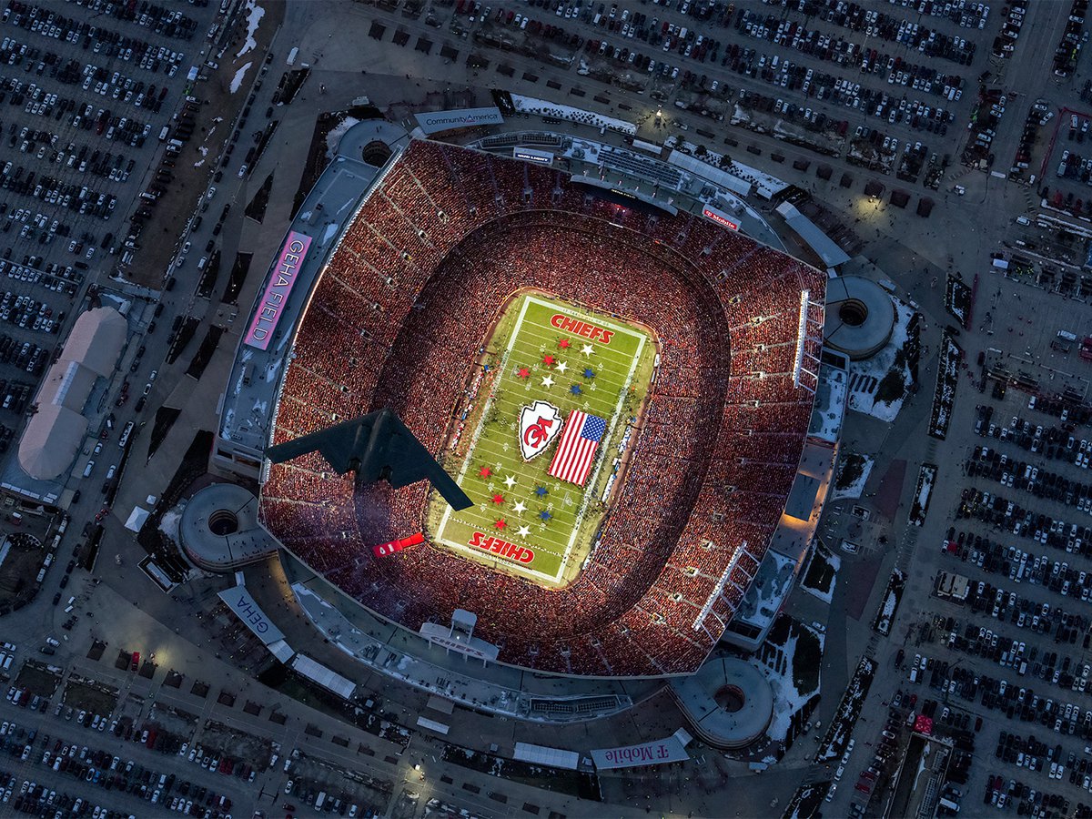 An aerial photograph capturing a B-2 stealth bomber soaring over GEHA Field during the 2025 AFC Championship Game, with a vibrant sea of red Kansas City Chiefs fans filling the stands and a large American flag displayed on the illuminated field below.