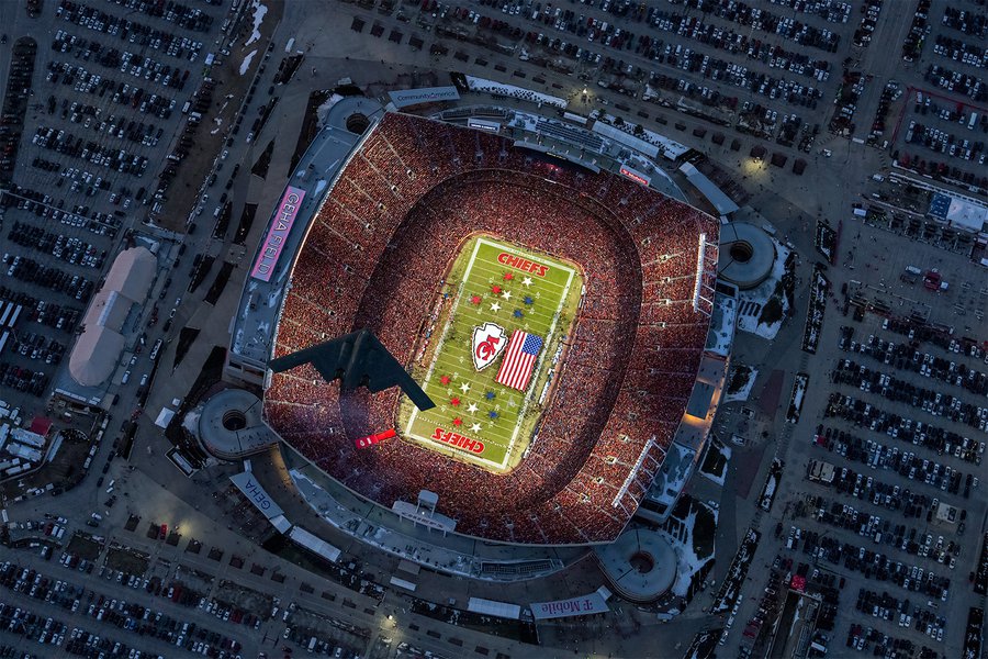 An aerial photograph capturing a B-2 stealth bomber soaring over GEHA Field during the 2025 AFC Championship Game, with a vibrant sea of red Kansas City Chiefs fans filling the stands and a large American flag displayed on the illuminated field below.